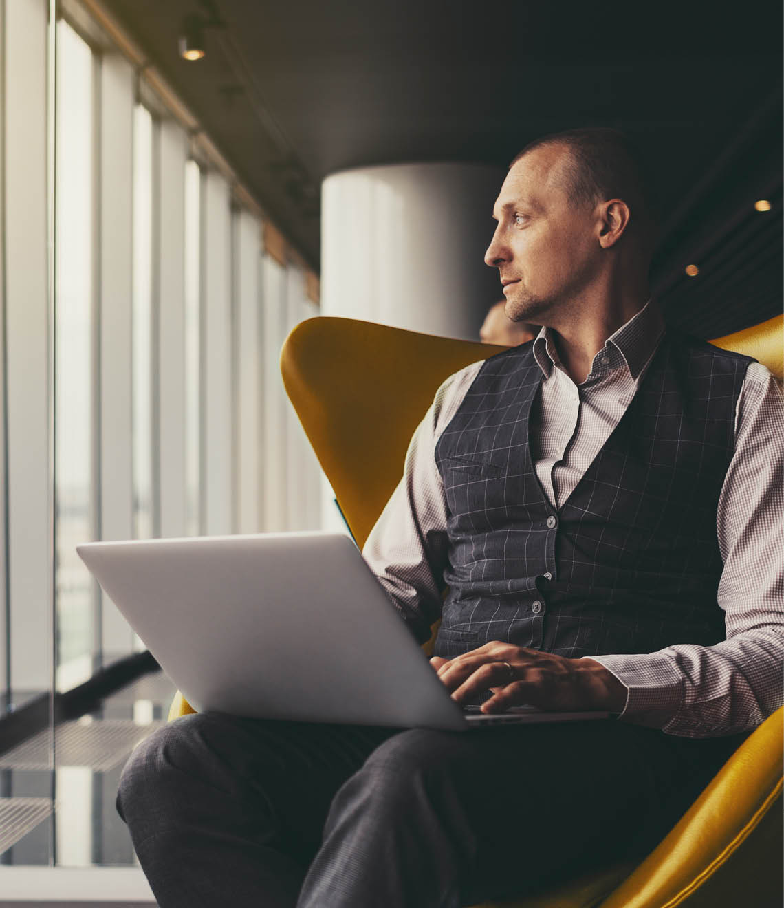 A confident relaxed successful mature man entrepreneur sitting with a laptop on an orange armchair and thoughtfully looking aside on an urban skyline outside the window; a copy space place on the left