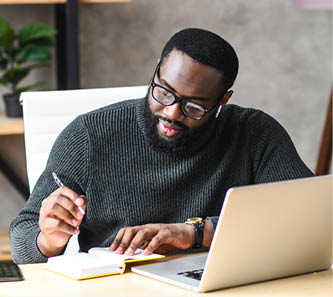 Smiling African-American guy sits at the desk in modern office, young black man in glasses is watching at laptop screen and writing notes in notebook
