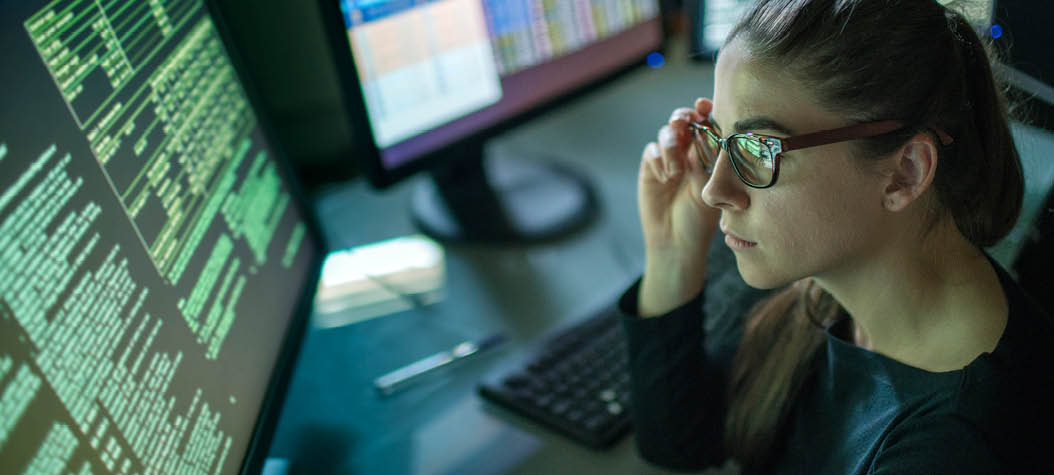 A young woman is seated at a desk surrounded by monitors displaying data, she is contemplating in this dark, moody office 
